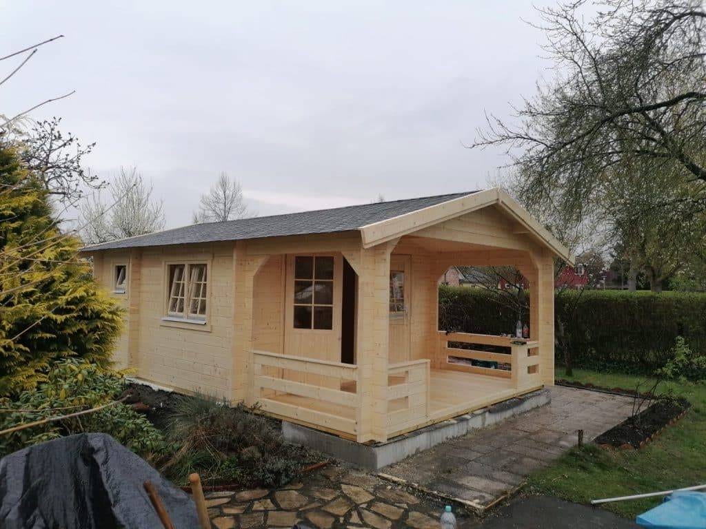 Das hölzerne Gartenhaus Modell Oslo mit angeschlossener offener Veranda befindet sich in der Aufbauphase und steht auf einer gepflasterten Fläche im Hinterhof eines Wohnhauses. Dieses herausragende Bauwerk steht prächtig unter einem bewölkten Himmel und unterstreicht seine einzigartigen Merkmale und die Liebe zum Detail in der Handwerkskunst.