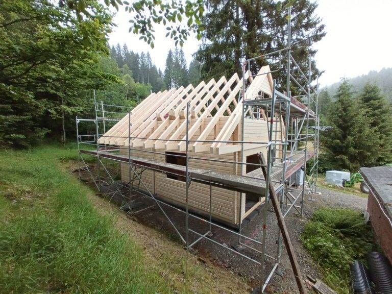 Ein im Bau befindliches Luxus-Holzhaus, umgeben von einem Gerüst. Der Dachstuhl besteht teilweise aus Holzbalken und die Wände sind gerade im Bau. Das Grundstück ähnelt einem charmanten Blockhaus, eingebettet in ein Waldgebiet, im Hintergrund sind Bäume und Grünflächen zu sehen.