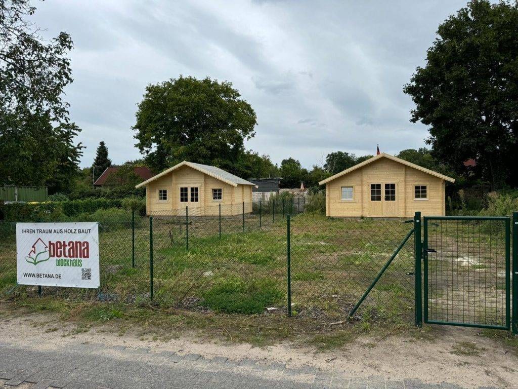 Zwei kleine Holzhäuser stehen in einem eingezäunten Bereich, umgeben von Grün. Auf dem Zaun hängt ein Schild mit der Aufschrift „Betana Blockhaus“ und einer Website-URL. Der Himmel ist bedeckt, und im Hintergrund stehen größere Bäume, was auf eine friedliche Atmosphäre hindeutet, die typisch für eine Kleingartenanlage wie Schlachtensee ist.
