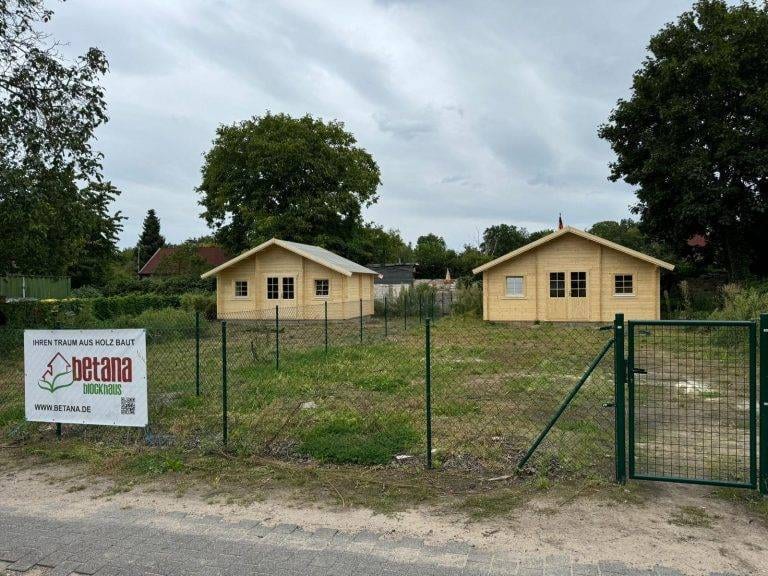 Zwei kleine Holzhäuser stehen in einem eingezäunten Bereich, umgeben von Grün. Auf dem Zaun hängt ein Schild mit der Aufschrift „Betana Blockhaus“ und einer Website-URL. Der Himmel ist bedeckt, und im Hintergrund stehen größere Bäume, was auf eine friedliche Atmosphäre hindeutet, die typisch für eine Kleingartenanlage wie Schlachtensee ist.