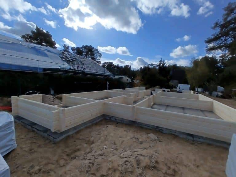Auf einer Baustelle wird der Holzrahmen eines Gebäudes im Bungalow-Stil auf einem Sandgrundstück zusammengebaut. Umgeben von verschiedenen Werkzeugen und Materialien ist im Hintergrund unter einem teilweise bewölkten Himmel eine Solaranlage zu sehen.