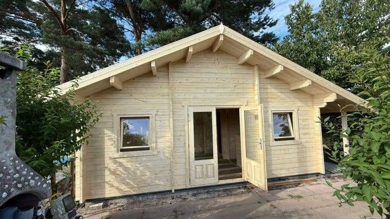 Eine kleine Holzhütte mit offener Eingangstür und zwei Fenstern, umgeben von Grün und Bäumen, ist wie geschaffen für Projekte. Das Schrägdach mit freiliegenden Balken sitzt auf einer Steinterrasse unter einem teilweise bewölkten Himmel und lädt zur Inspiration ein.