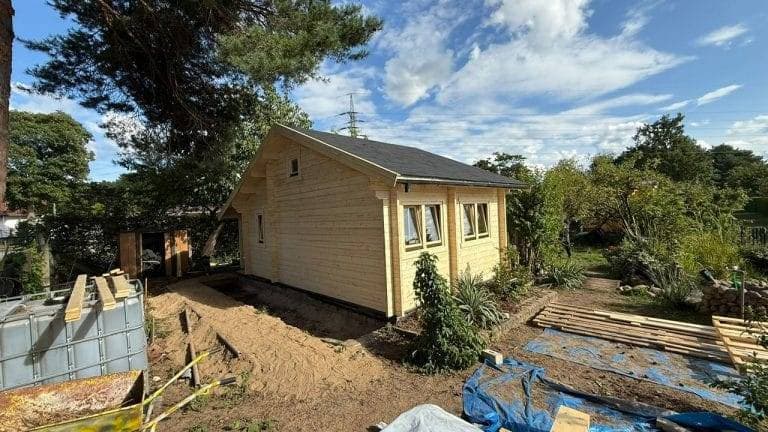 Ein kleines, neu gebautes Holzhaus mit einem dunklen Schindeldach ist von Erde, Baumaterialien und Grünpflanzen unter einem teilweise bewölkten Himmel umgeben. Im Hintergrund sind Bäume und Sträucher zu sehen.