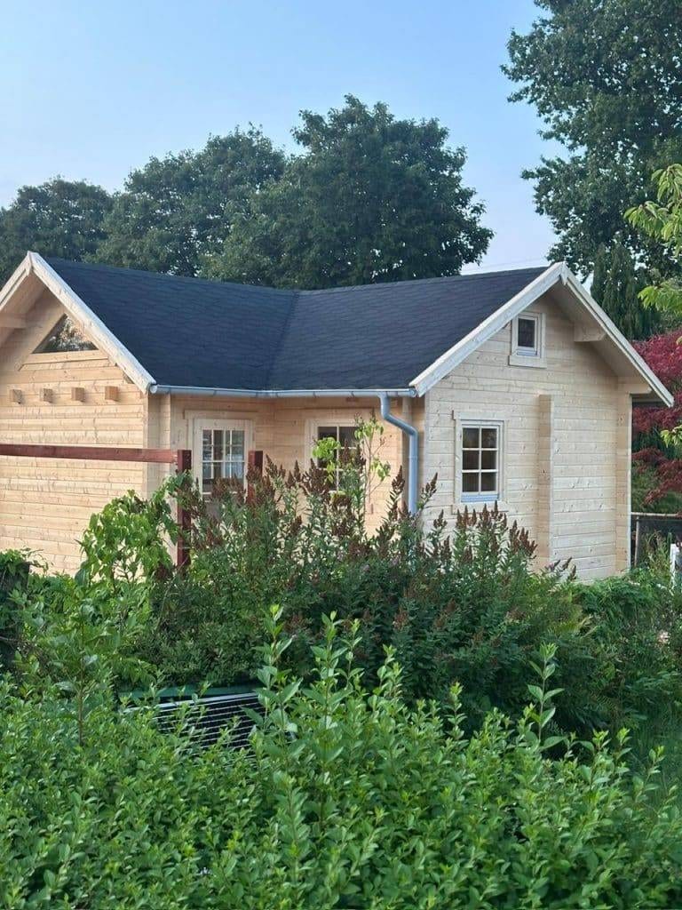 Ein kleines, helles Holzhaus mit dunklem Dach ist von grünen Sträuchern und Bäumen umgeben - ein idealer Rahmen für Projekte und kreative Aktivitäten im Freien. Weiß gerahmte Fenster und ein schlichtes Design vervollständigen diesen einladenden Rückzugsort im Garten.