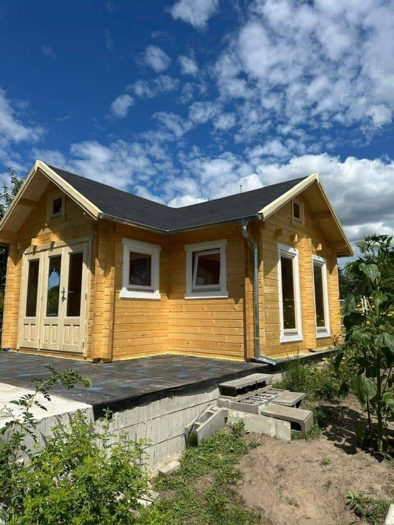 Ein kleines Holzhaus mit einem dunklen Schindeldach und mehreren weiß gerahmten Fenstern steht auf einem erhöhten Fundament. Der Himmel ist meist blau mit vereinzelten Wolken, während das Grün diesen von Projekten inspirierten Rückzugsort umgibt.
