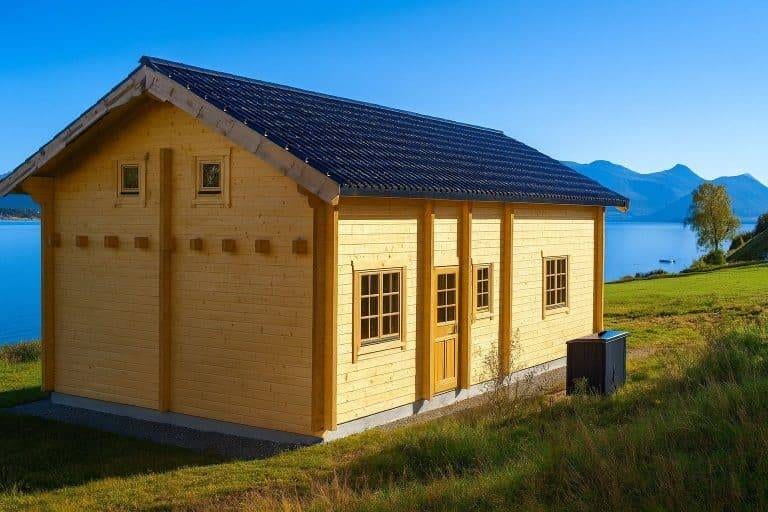 Eine kleine gelbe Holzhütte mit einem dunkelblauen Dach steht auf einem grasbewachsenen Hügel mit Blick auf einen See, mit Bergen und einem klaren blauen Himmel im Hintergrund.