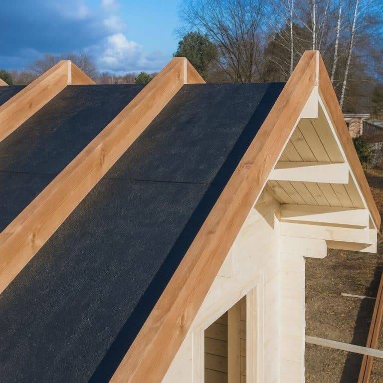 Eine Nahaufnahme eines im Bau befindlichen Holzhauses, die die Dachkonstruktion mit freiliegenden Holzbalken, schwarzer Dachunterlage und installierter Isolierung zeigt. Im Hintergrund sind Bäume und ein teilweise bewölkter Himmel zu sehen.