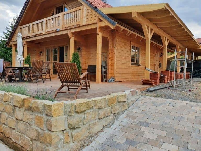 Ein Holzhaus mit überdachter Veranda und Balkon, Sitzgelegenheiten im Freien auf einer Holzterrasse, eine steinerne Stützmauer und Stapel von Dachziegeln neben einem gepflasterten Weg unter einem bewölkten Himmel.