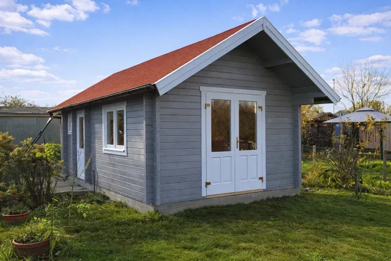 Blockhaus Modell Gardasee: Graues Holzhaus mit rotem Dach und weißen Fenstern im Garten.
