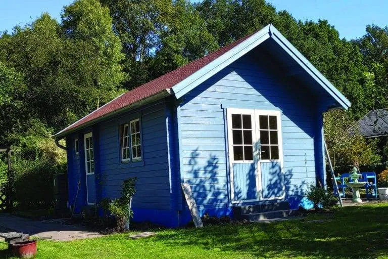 Abgebildet ist ein hellblaues Schlafbodenhaus aus Holz mit einem braunen, schrägen Dach. Es hat mehrere Fenster, eine weiße Tür mit Glasscheiben und steht auf einer Rasenfläche mit Bäumen im Hintergrund. Gartengeräte und eine kleine Leiter stehen in der Nähe der 24 m² großen Gartenlaube.