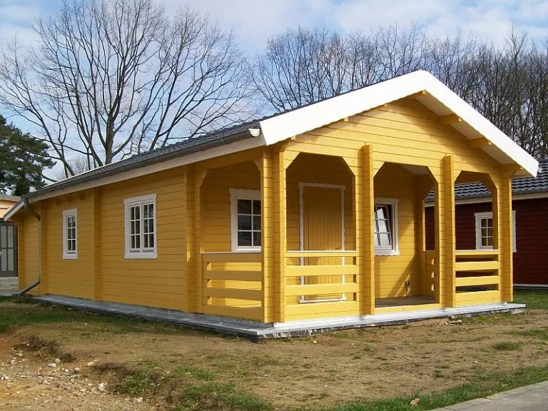 Ein neu errichtetes gelbes Holzhaus mit einer Veranda und kahlen Bäumen im Hintergrund.