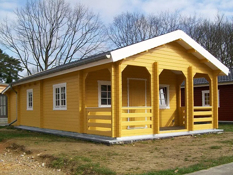Ein neu errichtetes gelbes Holzhaus mit einer Veranda und kahlen Bäumen im Hintergrund.