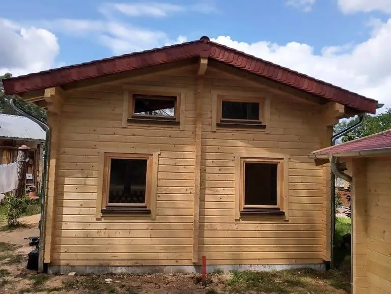 Ein kleines Ferienhaus aus hellem Holz, braun gerahmten Fenstern und einem roten Ziegeldach. Das Haus hat zwei rechteckige Fenster im unteren Stockwerk und zwei kleinere quadratische Fenster im oberen Stockwerk. Eine bezaubernde Veranda ziert die Vorderseite, perfekt, um den teilweise bewölkten Himmel zu genießen.
