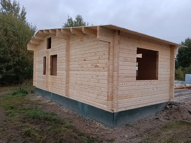 Ein im Bau befindliches Holzblockhaus präsentiert seine Sperrholzwände und Fensteröffnungen. Auf einem Fundament mit Gras und Bäumen im Hintergrund strahlt dieses Haus skandinavisches Flair vor dem bedeckten Himmel aus.