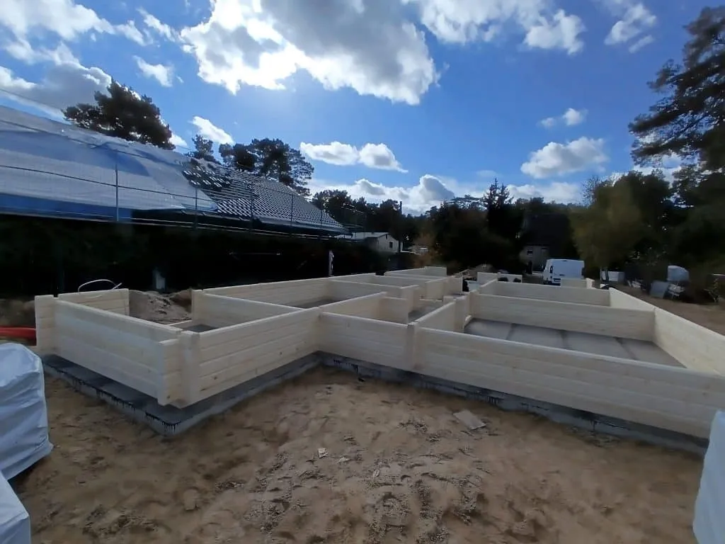 Auf einer Baustelle wird der Holzrahmen eines Gebäudes im Bungalow-Stil auf einem Sandgrundstück zusammengebaut. Umgeben von verschiedenen Werkzeugen und Materialien ist im Hintergrund unter einem teilweise bewölkten Himmel eine Solaranlage zu sehen.